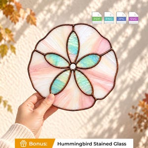 Puede incluir: Un atrapasoles de vitral circular con forma de dólar de arena, con segmentos iridiscentes en forma de pétalo de color verde azulado rodeados por paneles de vidrio blanco translúcido y rosa pálido, unidos por líneas de soldadura de metal oscuro.