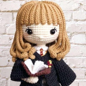 May include: A crocheted doll with light brown yarn hair, black eyes, and embroidered eyelashes. The doll wears a black robe over a white shirt with a red and yellow striped tie. It holds an open book with white pages and a brown cover.