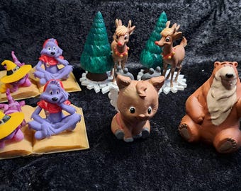 Könnte beinhalten: Eine Sammlung kleiner Kunststofffiguren, darunter zwei lila Wesen, die auf offenen Büchern mit gelben Hüten sitzen, zwei Rentiere neben grünen Tannenbäumen, ein kleines geflügeltes Wesen und eine große braune Bärenfigur.