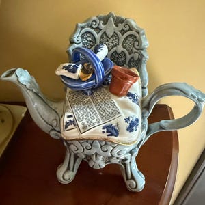 Peut inclure: Une théière décorative en céramique avec un corps en forme de chaise ornée bleu clair et blanc. Le couvercle est décoré d'un panier bleu miniature, d'un petit pot en terre cuite marron et d'un tissu blanc imprimé de texte noir et motifs bleus.