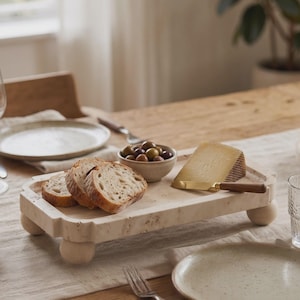 May include: A rectangular beige travertine serving board with four rounded wooden feet. The board holds three slices of crusty bread, a small ceramic bowl filled with green and black olives, a wedge of hard cheese, and a gold-toned cheese knife.