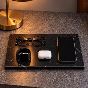Könnte beinhalten: Ein rechteckiges Tablett aus schwarzem Marmor mit weißer Maserung trägt eine Brille mit dunklem Rahmen, eine schwarze Smartwatch mit Silikonarmband, ein weißes Ladeetui für Ohrhörer und ein schwarzes Smartphone.