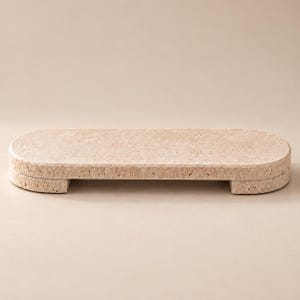 Pode incluir: Uma bandeja oval alongada feita de pedra travertino bege com uma superfície porosa e texturizada. A bandeja apresenta uma superfície superior plana e é apoiada por dois pés de bloco curtos integrados na parte inferior.