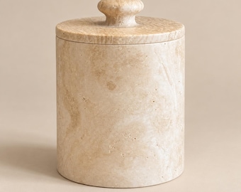 May include: A cylindrical beige travertine canister with a matching circular lid. The stone features a natural porous texture with swirling patterns in shades of tan and cream. A small, rounded knob sits centred on top of the flat lid.