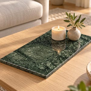 May include: A rectangular green marble tray with dark veining. A cylindrical white candle and a small, round, speckled beige ceramic vase containing a green leafy sprig are placed on the polished, smooth surface of the stone tray.