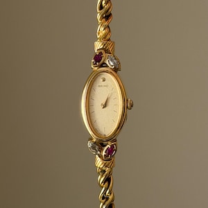 Puede incluir: Un reloj Seiko dorado con esfera ovalada y fondo color champán texturizado. El reloj cuenta con una correa de cadena trenzada y está adornado con piedras rojas y transparentes de corte marquesa en la parte superior e inferior de la caja.