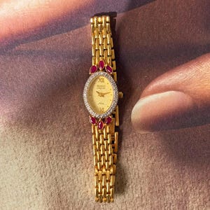 May include: A gold-tone wristwatch with an oval face, featuring a champagne dial with Roman numerals at the twelve and six o'clock positions. The bezel is encrusted with clear stones and accented by four marquise-cut red stones at the top and bottom.