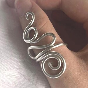May include: A silver-toned wire ring featuring a continuous, fluid design with multiple interconnected loops and swirls. The metal wire is shaped into an elongated, abstract pattern that wraps around the finger, showcasing a polished, smooth surface.
