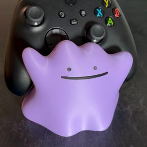 Könnte beinhalten: Ein schwarzer kabelloser Videospiel-Controller, der von einer violetten, amorphen, klecksartigen Figur mit zwei kleinen kreisförmigen Augen und einem dünnen, horizontalen Strich als Mund gehalten wird. Der Controller hat zwei Analogsticks und ein Steuerkreuz.