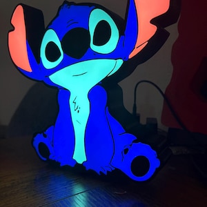 May include: A glowing LED lamp shaped like the character Stitch, featuring a vibrant blue body, a light cyan chest and face, and large ears with illuminated coral-pink inner sections. The lamp emits a bright, colorful light from its edges.