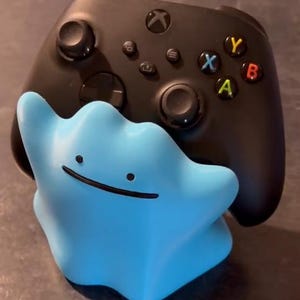 May include: A black wireless video game controller resting on a light blue, blob-shaped stand. The stand features a simple face with two small black dots for eyes and a thin, curved black line for a mouth, designed to hold the controller upright.