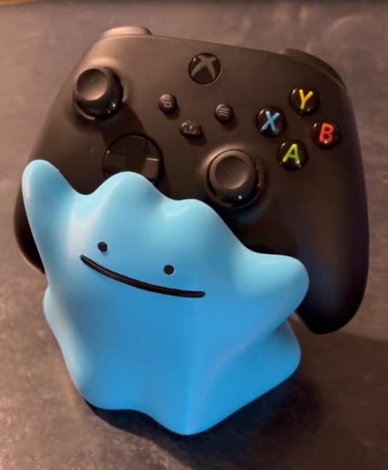 May include: A black wireless video game controller resting on a light blue, blob-shaped stand. The stand features a simple face with two small black dots for eyes and a thin, curved black line for a mouth, designed to hold the controller upright.