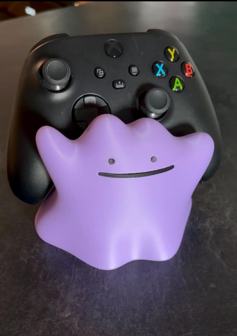 May include: A black wireless video game controller held by a purple, amorphous, blob-like figure with two small circular eyes and a thin, horizontal line for a mouth. The controller features two analog sticks, a directional pad, and four colored buttons.