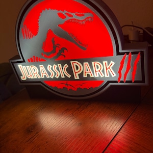 Può includere: Un'insegna retroilluminata con il logo di Jurassic Park, uno sfondo circolare rosso, una sagoma nera di dinosauro e la scritta JURASSIC PARK in bianco. L'insegna presenta tre graffi rossi sul lato destro e una cornice nera.