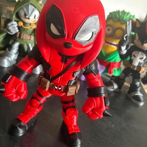 Può includere: Un action figure rosso e nero che raffigura un personaggio con cappuccio, grandi occhi bianchi e una cintura multiuso con fibbia circolare. Il personaggio è in una posa dinamica con i pugni chiusi, circondato da altre statuette sullo sfondo.