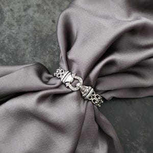 May include: A silver-toned metal scarf ring featuring a central horseshoe-shaped link with embedded clear stones, flanked by twisted rope-style accents and rectangular end pieces, securing a smooth, draped grey satin fabric.