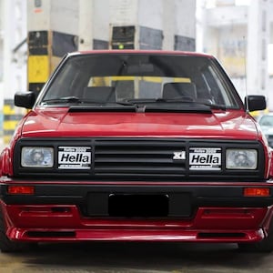 Puede incluir: Un coche rojo visto de frente con una parrilla negra que integra dos luces rectangulares Hella Rallye 3000. El vehículo cuenta con faros cuadrados, un parachoques negro y un pequeño emblema xi situado en el centro de la parrilla.