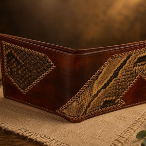 May include: A brown leather bifold wallet featuring two triangular inlays of textured, snakeskin-patterned material. The inlays are secured with visible, light-coloured cross-stitching along the edges, contrasting with the smooth, dark brown leather.