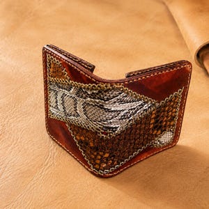 May include: A bifold wallet crafted from brown leather featuring a patchwork design. The exterior displays contrasting panels of textured snakeskin in shades of grey, white, and dark brown, accented with visible stitching along the edges.