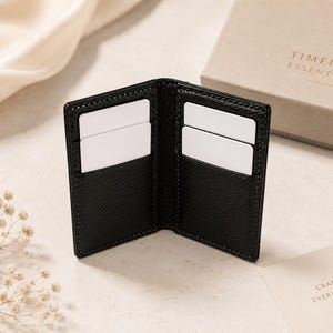 May include: A black textured leather card holder with four interior slots containing white blank cards. The card holder is positioned near a beige gift box with the text TIMELESS ESSENTIALS and a card with the text CRAFTED FOR EVERYDAY LUXURY.