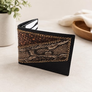 May include: A bifold wallet featuring a black leather frame with a central panel of brown snakeskin-patterned material and a top section of dark brown textured leather, all joined by visible tan cross-stitching along the edges of the panels.