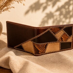 May include: A bifold wallet crafted from dark brown leather and tan suede, featuring a geometric patchwork design with textured snakeskin-patterned panels. The exterior is accented with visible light-coloured cross-stitching along the seams.