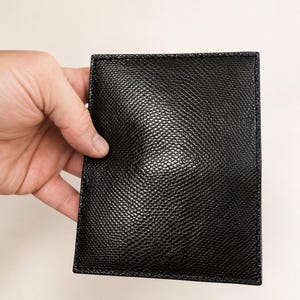 Op de afbeelding: Een rechthoekige zwarte portemonnee met een getextureerd, generfd oppervlak. De portemonnee heeft strakke, gestikte randen en een gestructureerd, plat ontwerp. Het materiaal heeft een lichte glans die het licht reflecteert in het midden.