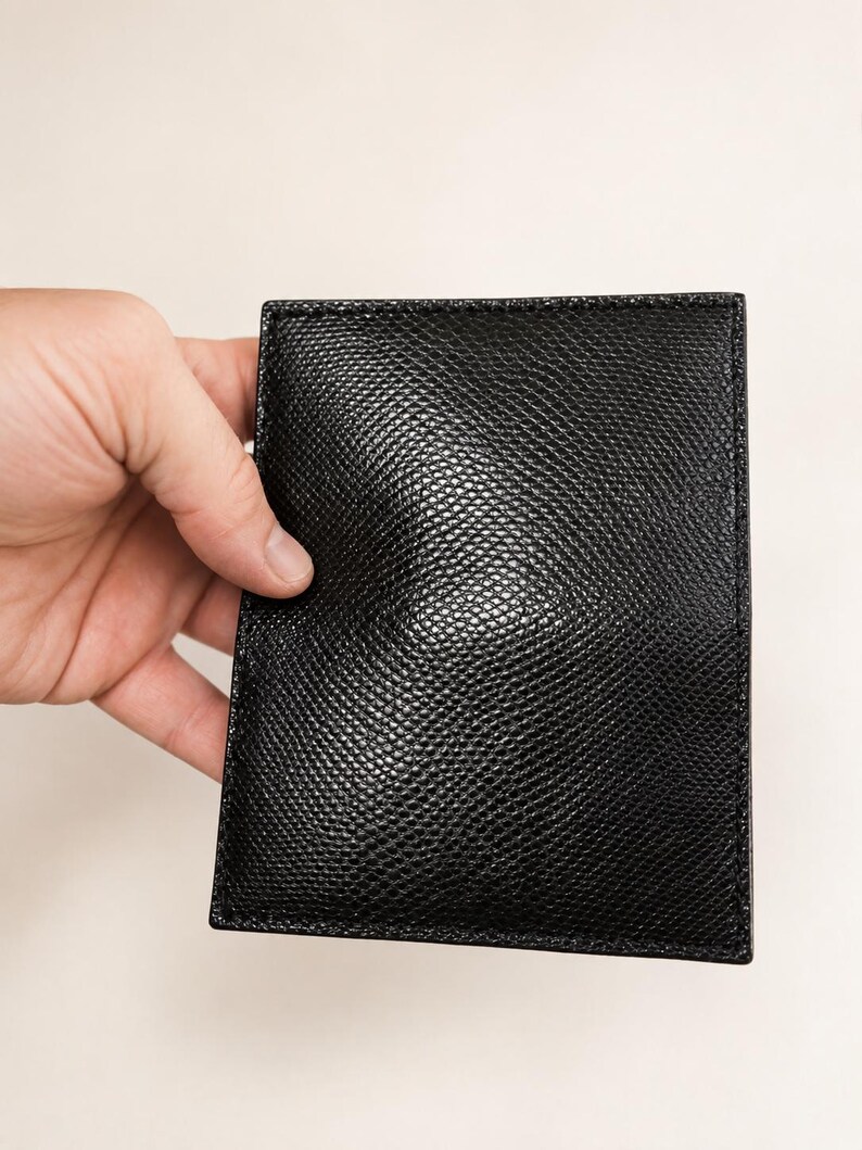 Op de afbeelding: Een rechthoekige zwarte portemonnee met een getextureerd, generfd oppervlak. De portemonnee heeft strakke, gestikte randen en een gestructureerd, plat ontwerp. Het materiaal heeft een lichte glans die het licht reflecteert in het midden.