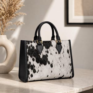 May include: A structured handbag featuring a black and white cowhide print on the front panel. The bag includes black side panels, two top handles, and gold-toned hardware attachments connecting the handles to the main body of the bag.