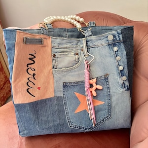 Puede incluir: Un bolso tote de mezclilla tipo patchwork con un panel rosa claro que tiene el texto negro "merci" y un pequeño corazón rojo. El bolso incluye un asa de cuentas de perlas blancas, una estrella naranja grande en un bolsillo y un dije de perro globo rosa.