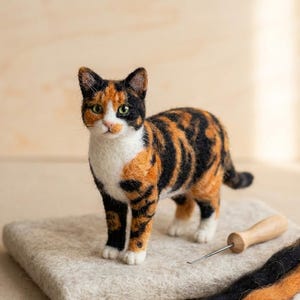 May include: A needle-felted calico cat figurine with black, orange, and white fur, green eyes, and a pink nose. The cat stands on a rectangular felt pad next to a wooden-handled felting needle and three strands of loose black, orange, and white wool.