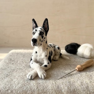 May include: A needle-felted Great Dane figurine with a white coat and black spots, depicted in a lying position with front paws crossed. The sculpture features upright ears, a black nose, and dark eyes, accompanied by a wooden felting needle tool.