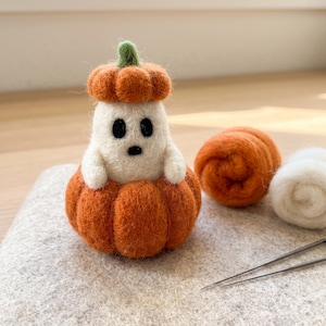 Puede incluir: Una figura de fantasma de lana fieltrada sentada dentro de una calabaza naranja texturizada, con un pequeño sombrero de calabaza naranja con tallo verde. El fantasma tiene ojos y boca de fieltro negro. Junto a él, dos rollos de lana y dos agujas.