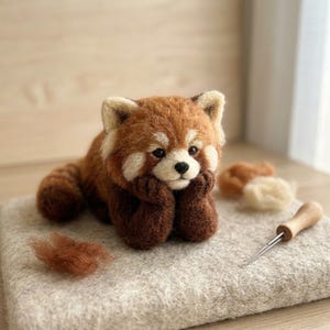 May include: A needle-felted red panda figurine with reddish-brown and white fur, dark brown limbs, and black eyes. The panda rests its chin on its front paws. Nearby are loose tufts of wool and a wooden-handled felting needle on a felt pad.