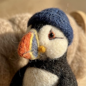 May include: A needle-felted puffin figurine wearing a dark blue knit cap. The puffin has a white face, black body, and a multi-colored beak featuring orange, yellow, and blue sections. The bird has a small black eye and a textured wool finish.