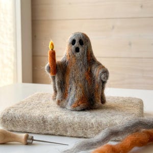 Puede incluir: Una figura de fantasma hecha con fieltro de aguja a partir de fibras de lana gris y naranja. El fantasma presenta dos orificios oscuros para los ojos y una pequeña abertura bucal, y sostiene una vela naranja brillante con llama amarilla.