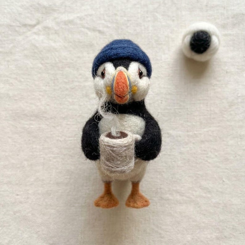 May include: A needle-felted puffin figurine wearing a dark blue knit hat, holding a small beige mug with a textured surface. The puffin has a white face, black wings, an orange beak, and orange feet. A separate small white felted ball is nearby.