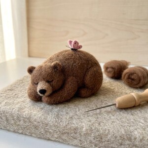May include: A needle-felted brown bear figurine in a sleeping position with a small pink butterfly perched on its back. The bear has a dark nose and closed eyes. Nearby are two small brown wool rolls and a wooden needle felting tool.