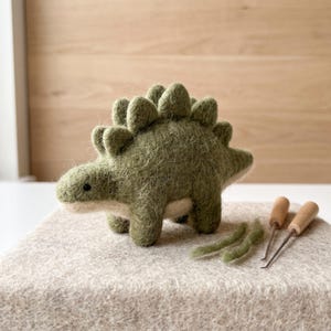 May include: A needle-felted olive green stegosaurus dinosaur figurine with a textured, fuzzy surface, a light beige underbelly, and small black bead eyes, accompanied by two metal felting needles and loose green wool fibers.