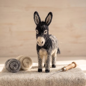May include: A needle-felted donkey figurine with black and grey wool fibres, featuring a light-coloured muzzle and upright ears. Beside the donkey are two small rolls of grey and cream wool roving and a metal felting needle with a wooden handle.