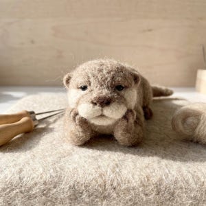 May include: A needle-felted sculpture of an otter with light brown and cream-coloured wool fibres. The otter is lying down with its front paws tucked under its chin, featuring dark eyes and a dark brown nose, crafted with a textured, fuzzy finish.