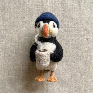 May include: A needle-felted puffin figurine wearing a dark blue knit-style cap. The puffin has a black and white body, an orange beak with yellow accents, and orange feet. It holds a small, light beige mug with wisps of white felt steam.