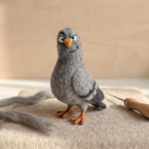 May include: A needle-felted grey pigeon figurine with a textured wool finish, featuring a small orange beak, orange feet, and a distinctively grumpy facial expression with raised eyebrows and wide eyes, standing upright on a light-coloured felt pad.