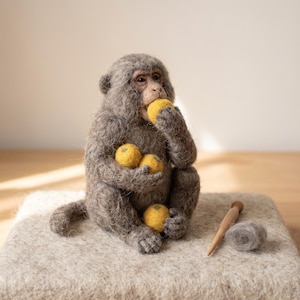 May include: A needle-felted gray monkey figurine with a textured wool finish, holding and eating small yellow round fruits. Beside the monkey sits a wooden felting tool and a small tuft of gray wool fiber, all crafted with detailed realistic features.