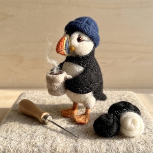 May include: A needle-felted puffin figurine wearing a dark blue knit cap, holding a small beige mug with wisps of white wool steam. The puffin has a black and white body with orange feet and a multi-colored beak. A metal felting needle and wool balls sit nearby.