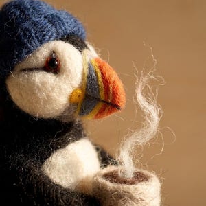 May include: A needle-felted puffin figurine wearing a dark blue knit hat. The puffin has a white face, black body, and a multi-colored beak with orange, yellow, and blue stripes. It holds a small beige cup with wisps of white fiber steam rising.