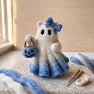 Puede incluir: Una figura de fantasma de lana fieltrada en blanco con una base en degradado azul, con un lazo azul en la cabeza y sosteniendo un pequeño cubo de calabaza azul. El fantasma tiene dos ojos negros y está junto a fibras de lana azules y blancas.