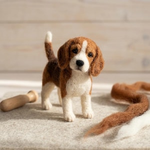 Könnte beinhalten: Eine nadelgefilzte Beagle-Figur mit einem dreifarbigen Fell aus brauner, schwarzer und weißer Wolle. Der kleine Hund steht mit aufrechtem Schwanz und zeigt Schlappohren, dunkle Augen und eine schwarze Nase in einer texturierten Faseroberfläche.