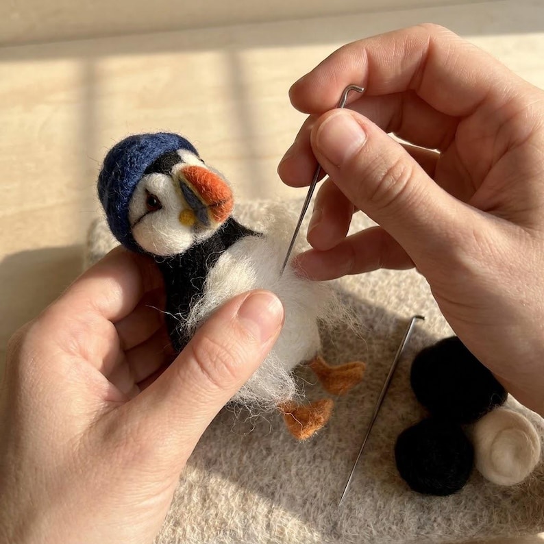 May include: A needle-felted puffin figurine with a dark blue hat, black and white body, and orange beak and feet. A metal felting needle is being used to shape the white wool fibers on the chest, with additional wool roving nearby.