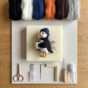 May include: A needle felting kit featuring a puffin figure wearing a blue hat, positioned on a foam block. Included are six bundles of wool roving in black, white, blue, orange, brown, and gray, plus scissors, needles, a tool handle, and a spray bottle.